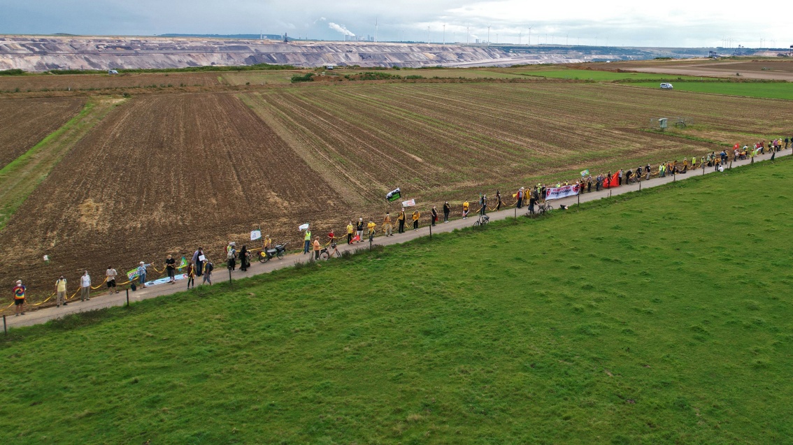 Vor dem Hintergrund eines Tagebaugeländes und großer Felder haben sich auf einem schmalen Feldweg zahlreiche Menschen mit Bannern zusammengefunden, um eine Menschenkette zu bilden.