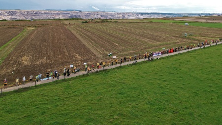 Vor dem Hintergrund eines Tagebaugeländes und großer Felder haben sich auf einem schmalen Feldweg zahlreiche Menschen mit Bannern zusammengefunden, um eine Menschenkette zu bilden.