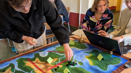Ein handbemaltes Tuch mit eineRr Landkarte, die durch zwei rote Dekobändern in Bereiche unterteilt ist, ein Spieler greift nach Spielsteinen aus farbigen Klemmbausteinen.