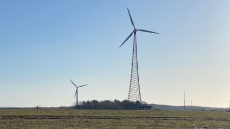 Zwei Windkraftwerke auf einer großen Wiese, im Hintergrund ein Wäldchen