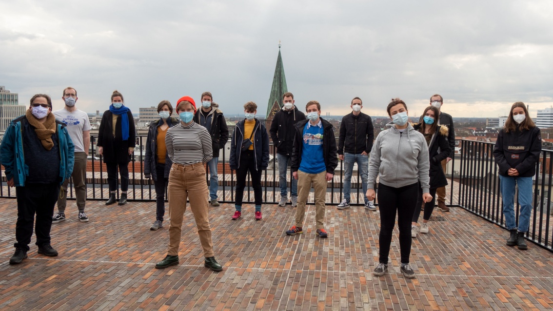 Eine Gruppe von vierzehn jungen Menschen mit Mundnasenschutz steht auf einem Dach über Bremen; sie stehen in coronakonformem Abstand voneinander entfernt und schauen selbstbewusst in die Kamera.