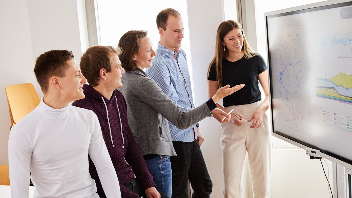 In einem Besprechungsraum schauen fünf junge Personen auf einen großen Monitor, auf dem eine Netzkarte Deutschlands neben einem Diagramm mit gestapelten Kurvenverläufen zu erkennen ist. Die mittlere Person, eine Frau, deutet mit nach oben geöffneter Handfläche auf den Monitor.