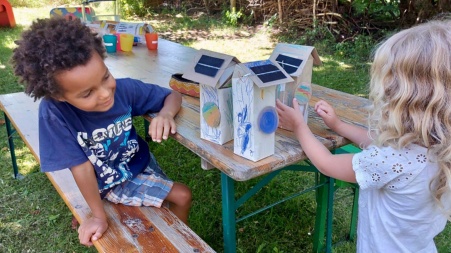 Zwei Kindergartenkinder an einem Tisch im Freien, sie basteln mit Solarmodulen, die an Kartonhäuschen befestigt sind.