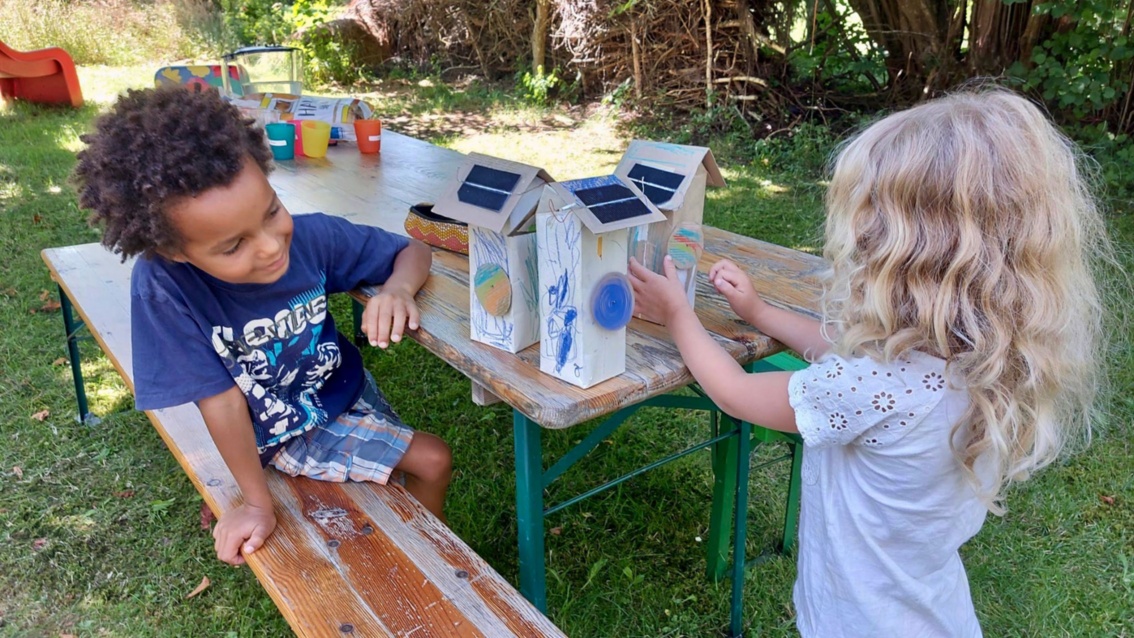 Zwei Kindergartenkinder an einem Tisch im Freien, sie basteln mit Solarmodulen, die an Kartonhäuschen befestigt sind.