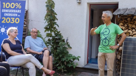 Ein Mensch referiert vor einer Haustür, weiterhin sind Plakate und ein PV-Modul im Bild; zwei Nachbar:innen sitzen daneben und hören zu.