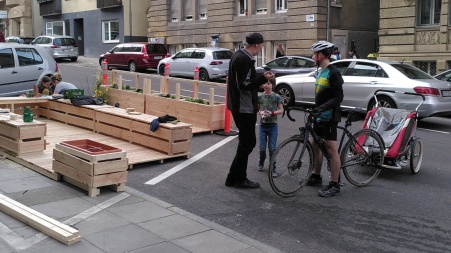 Ein junger Mann und ein Kind stehen neben einem im Aufbau befindlichen Parklet, daneben steht ein Radfahrer mit Helm.