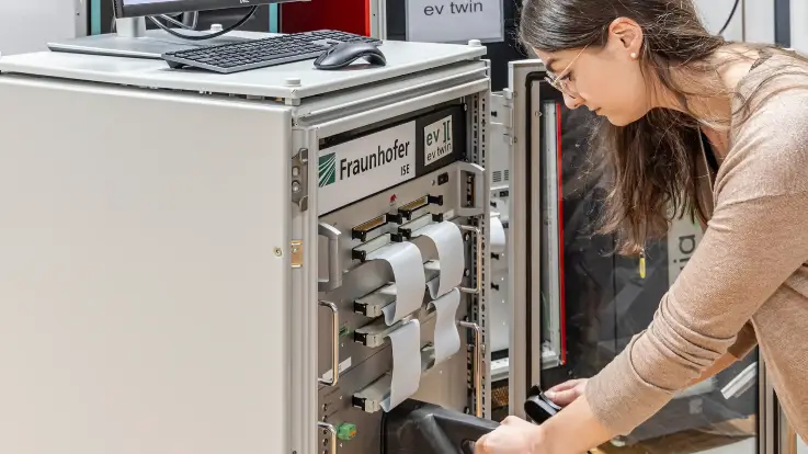 Eine junge Frau in einem Labor des Fraunhofer-Instituts, von technischen Geräten umgeben; sie steckt einen Stecker in eine Ladesäule