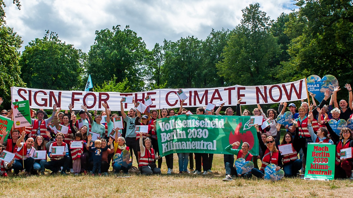 Eine Gruppe Menschen in einem Park; sie halten Banner mit der Aufschrift  «Sign for theclimate now» und «Berlin bis 2030 klimaneutral machen».