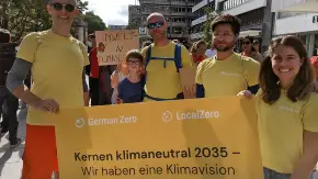 Ver jüngere Personen, eine von ihnen wohl Vater eines Kindes, das neben ihm steht, in gelben GermanZero-T-Shirts, halten ein gelbes Plakat mit der Aufschrift German Zero - LocalZero - Kernen klimaneutral 2035 - Wir haben eine Klimavision