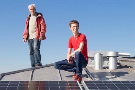 Auf einem Blechdach sitzt ein junger Mann vor Solarpanelen in der Hocke, im Hntergrund steht ein älterer Herr.